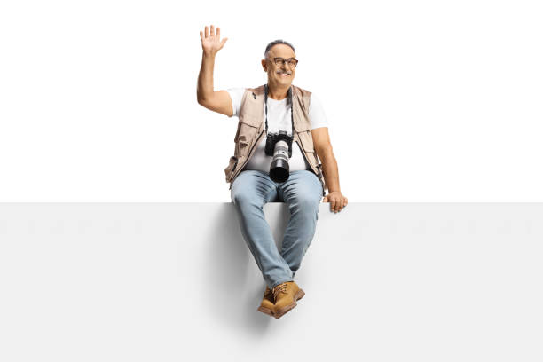 Man with a camera sitting on a blank panel and waving at camera isolated on white background