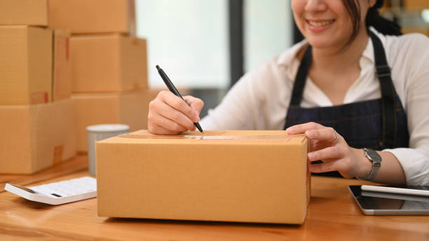 istock Female small business entrepreneur writing address on cardboard box while preparing parcel boxes of product in home office. 1415335194