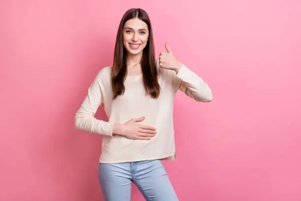 Portrait of attractive cheerful brown-haired girl touching belly showing thumbup isolated over pink pastel color background.