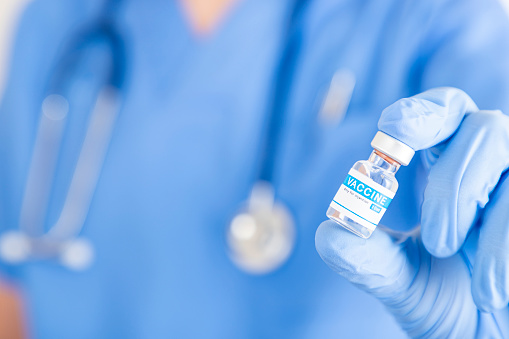 Front closeup view of an unrecognizable nurse wearing surgical gloves holding a vaccine vial at the hospital. Predominant color is blue. Selective focus is on the nurse hand.