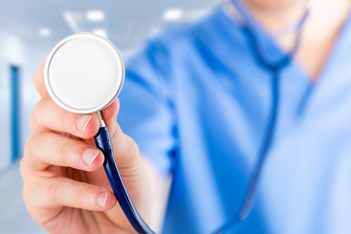 Front closeup view of an unrecognizable male nurse in uniform at the hospital holding a stethoscope and pointing it at camera. Predominant color is blue. Selective focus is on the nurse hand.