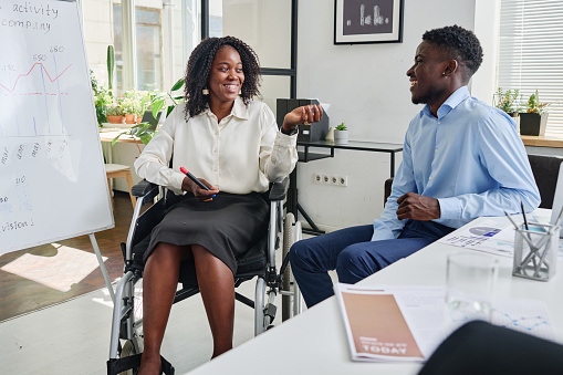African businesswoman sitting on wheelchair near the flipchart and discussing business strategy with her partner at office