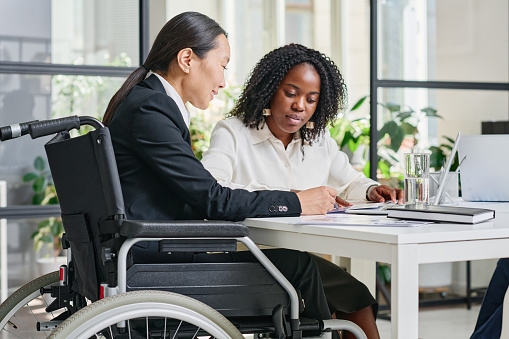 Asian businesswoman in wheelchair discussing business contract with her colleague at table at office