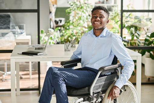 Portrait of African businessman with disability sitting in wheelchair and smiling at camera during his work at office