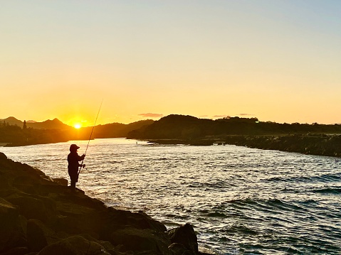 Horizontal seascape of sunset over unrecognizable fisherman at promenade rocks of calm river with coastline horizon at Brunswick Heads near Byron Bay Australia