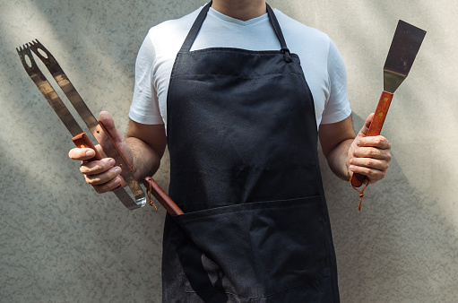 Un hombre con delantal de chef negro, sosteniendo herramientas de barbacoa: pinzas de barbacoa, espátula. photo
