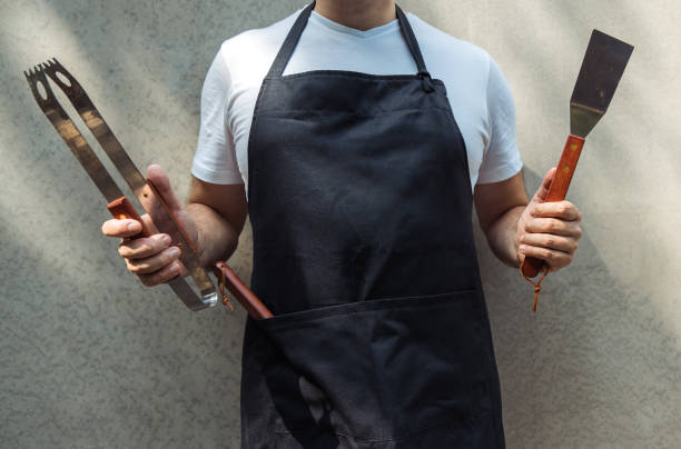 un uomo che indossa un grembiule nero da chef, con in mano strumenti da barbecue: pinze per barbecue, spatola. - grembiule foto e immagini stock