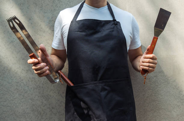 A man wearing black chef's apron, holding barbecue tools: bbq tongs, spatula. A man wearing black chef's apron, holding barbecue tools: bbq tongs, spatula. apron stock pictures, royalty-free photos & images