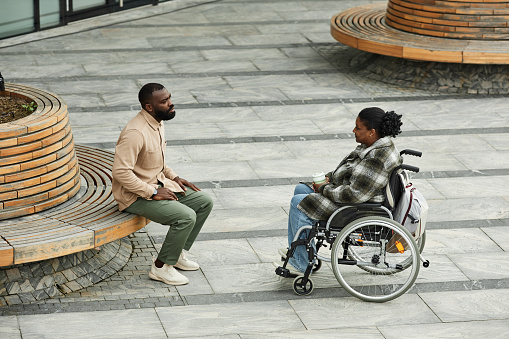 Wide angle side view at adult black couple with woman in wheelchair chatting outdoors in city setting and enjoying coffee, copy space