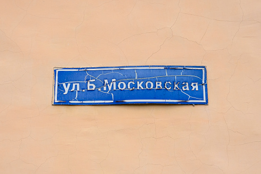 Weathered blue street name sign on cracked orange wall. The Russian sign says B. Moscovskaya st.