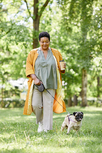 Vertical full length portrait of smiling black woman walking cute dog in park and enjoying coffee