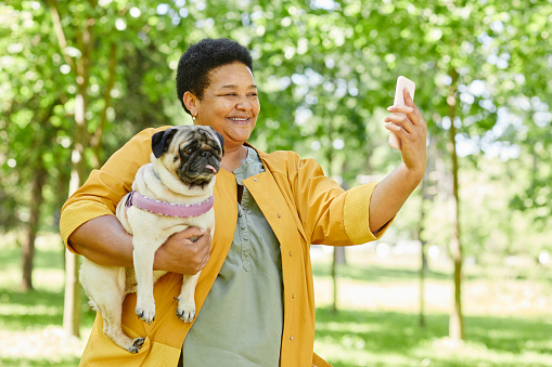 Waist up portrait of mature black woman taking selfie photo with dog in park and smiling happily