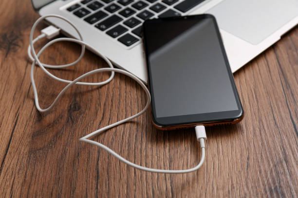 istock Smartphone connected with charge cable to laptop on wooden table, closeup. Space for text 1415359074