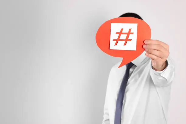 Photo of Man holding paper speech bubble with hashtag symbol in front of his face on light background. Space for text