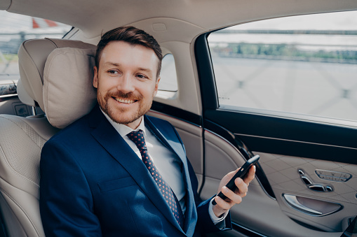 Cheerful young corporate executive in elegant expensive tuxedo rides in luxury car with company driver to financial meeting while checking market stock prices and indices on his cellphone