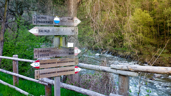 Wooden trail sign with arrows indicating the direction of different hikes, in the background flows the river the Passer.  Hike directions to Saltaus and Merano in northern Italy.
