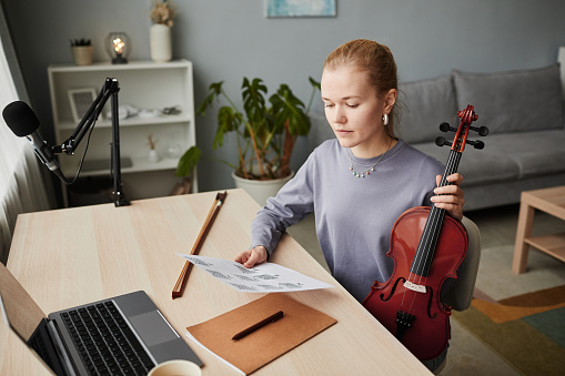 High angle portrait of blonde woman playing violin at home and looking at music sheets while composing, copy space