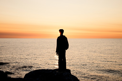 Silhouette of a young man standing in front of the sea at sunset.