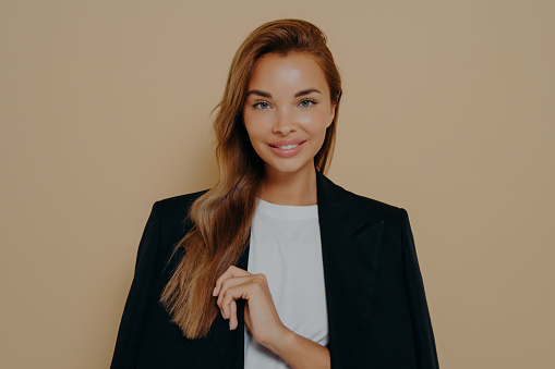 Friendly smiling brunette businesswoman with loose long hair, wears formal black jacket over white blouse, poses against beige background with blank space, happy to meet with business partners