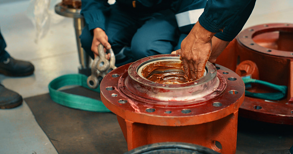 Male mechanics greasing a metal machine part in the workshop and fixing a mechanism for transmitting torque. Handyman coating or using lubrication on a steel gear or mechanical device