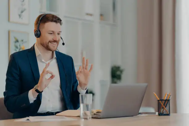 Photo of Career and distance job concept. Pleased bearded man corporate worker dressed formally gestures okay sign at webcam of laptop uses headset for online calling poses at desktop gains new knowledge