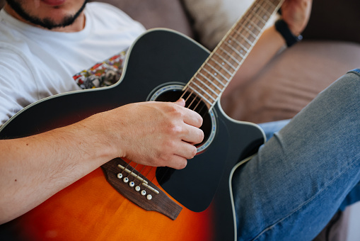 Close up portrait of young man playing acoustic guitar at home sitting on the gray couch, sofa in the living room. New hobby. Selective focus.