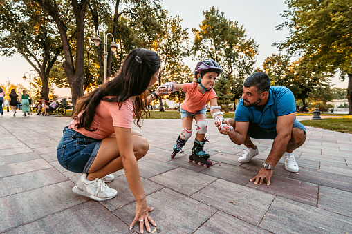 Young father and mother teaching their daughter how to roller skate in the park, on a sunny day.