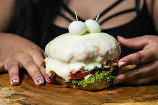 Colombian hamburger dipped in melted cheese, served on a wooden tray or plate, about to be eaten by a Colombian Latin girl. girl holding a hamburger with both hands.