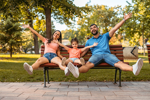 Young father and mother sitting on a park bench with their daughter, on a sunny day. Having fun.