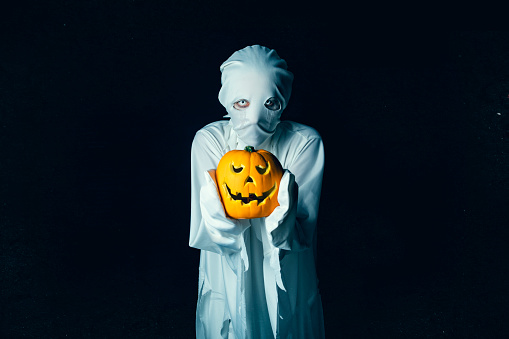 A creepy person dressed as a ghost holding a jack o' lantern on a black background with lots of copy space.