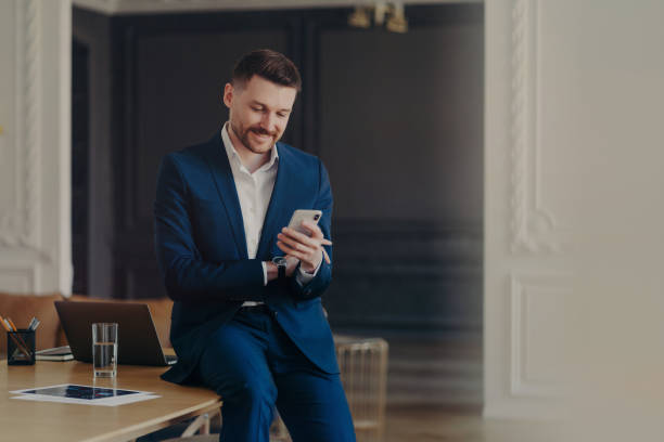 Happy handsome businessman looking at mobile phone while sitting on office desk