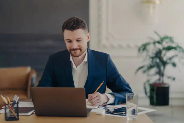 Photo of Indoor shot of handsome male office worker rewrites information from notepad in diary sits at desk dressed in formal clothes spends time for searching info. Businessman takes notes does paper work