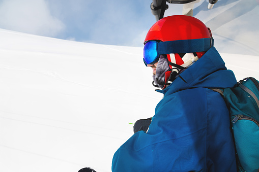 Portrait of a professionally dressed male skier in profile sitting in an open cabin of a ski lift waiting to ride down a slope against a blue sky and a snowy slope.