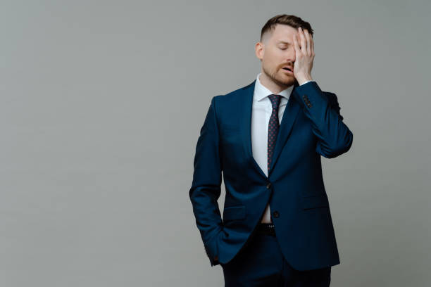 Upset man in suit feeling depressed of business failure Young worried man manager in formal wear feeling frustrated and upset about business failure, holding hand on face and keeping eyes closed while standing against grey background, copy space for text worried man standing stock pictures, royalty-free photos & images