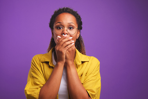 Frightened young ethnic African American woman covers mouth with hands experiencing negative emotions restrains himself so as not to scream and wants to avoid panic stands on purple background