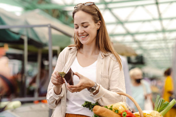 Smart food shopping A smiling young woman is holding a basket full of groceries and counting money financial wellbeing stock pictures, royalty-free photos & images