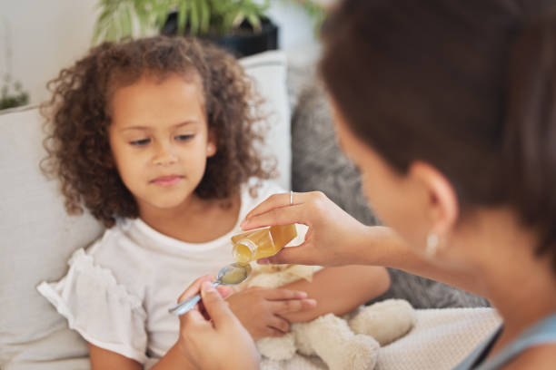 istock Sick daughter taking medicine as a treatment, cure or remedy while lying on the sofa for health and recovery at home. Ill girl being given medication with a spoon by her loving and caring mother 1415397120