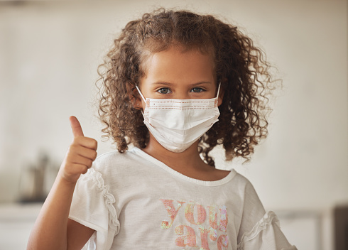 Little girl wearing a protective face mask with thumbs up gesture showing that she has protection or caution against disease, illness and virus. Confident child with good, happy and like emoji sign