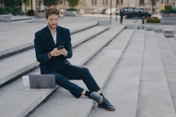 istock Young confident man in stylish suit using smartphone while sitting on steps outside with laptop 1415398635