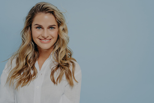 Attractive blonde woman in white shirt smiling and looking straight at camera with toothy smile and being calm at same time, posing alone next against blue background with copy space for text