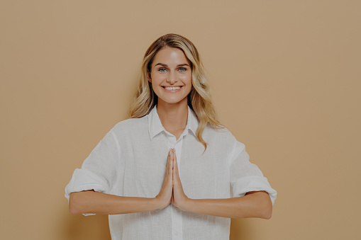 Happy cute blond feamle in white shirt pressing palms together in pray, namaste or saying please gesture, smiling gratefuly and thanking for help, isolated over beige studio background