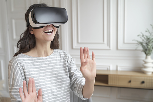 Young excited woman wearing 3d goggles interacting with augmented world, amazed female in VR headset playing games or making purchases in virtual reality store while spending leisure time at home