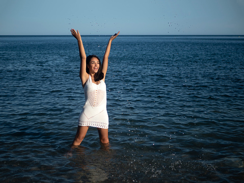Cheerful young ethnic female tourist with long dark hair in crochet dress smiling brightly and looking away while standing in shallow sea and splashing water on sunny day
