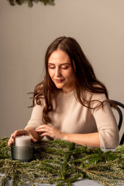 Young woman is sitting at table and looking at an unlit candle in middle of composition of Christmas tree branches. Close-up. Selective focus. stock photo