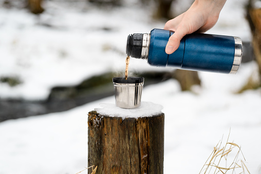 Male hand pouring hot tea from a thermos into a mug, close-up. Hot drink with steam in winter. Winter hiking concept, camping, travel.