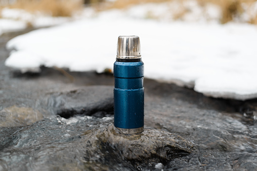 Blue vacuum thermos close-up outside in winter. Camping thermos standing on a stone in a winter stream. Adventure concept, winter hike, travel.