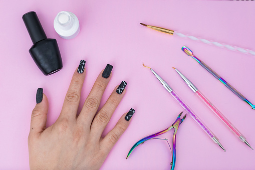 hand of a Latin girl, surrounded by basic elements for a manicure, placed on a pink background. black and white nail polish, colored brushes, cuticle pusher and cuticle nippers. sell of articles