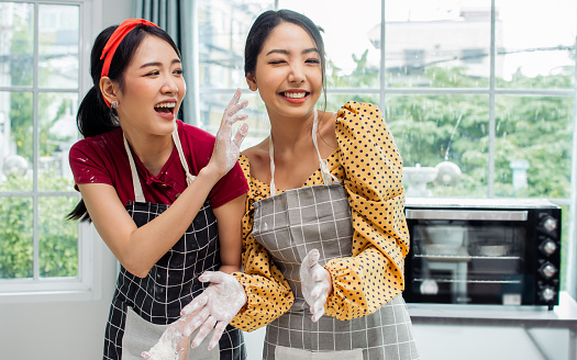 Two beautiful Asian women cooking food, making bread for breakfast and bakery, teasing, playing together with happiness, fun while standing in kitchen at home on weekend. Hobby and Lifestyle Concept
