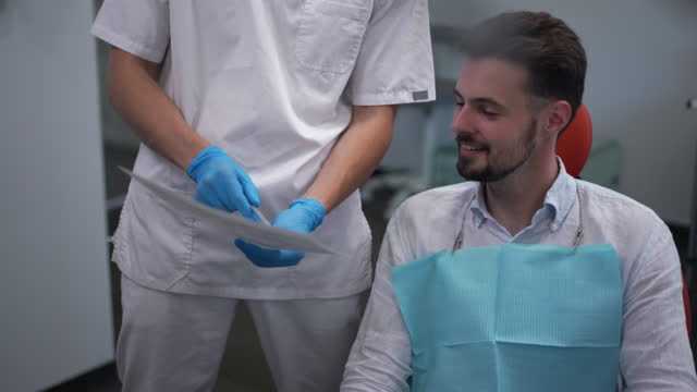 Unrecognizable dentist showing X-ray to patient sitting in dental chair smiling. Professional Caucasian male doctor planning treatment with man in medical clinic. Slow motion.