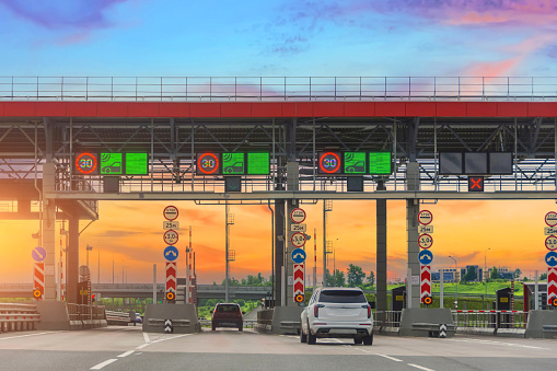 View with cars at the entrance to the toll road, limited by the barrier. Cashless payment transponder, speed limit signs. Sunset in the sky background